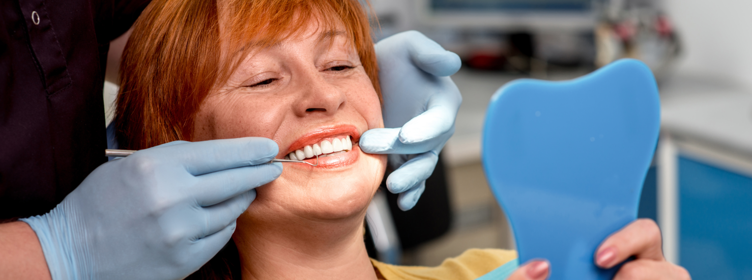 Eine Frau wird von einem Zahnarzt behandelt und lächelt in einen Spiegel.